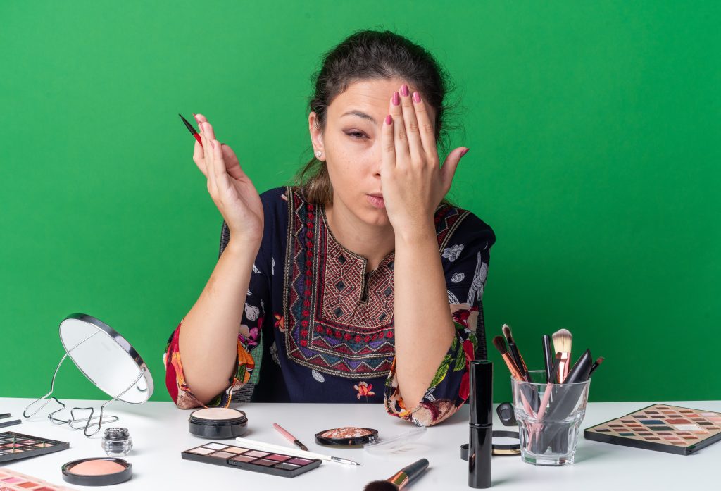 aching-young-brunette-girl-sitting-table-with-makeup-tools-holding-eyeliner-putting-hand-her-eye