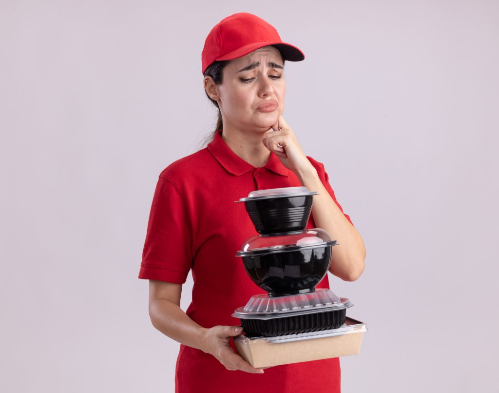frowning-young-delivery-woman-uniform-cap-holding-looking-paper-food-package-food-containers-touching-chin-isolated-white-wall-with-copy-space